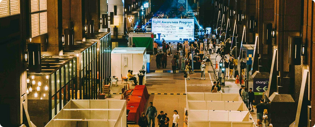 People walk through an indoor market with vendor stalls and a Sight Awareness Program sign.